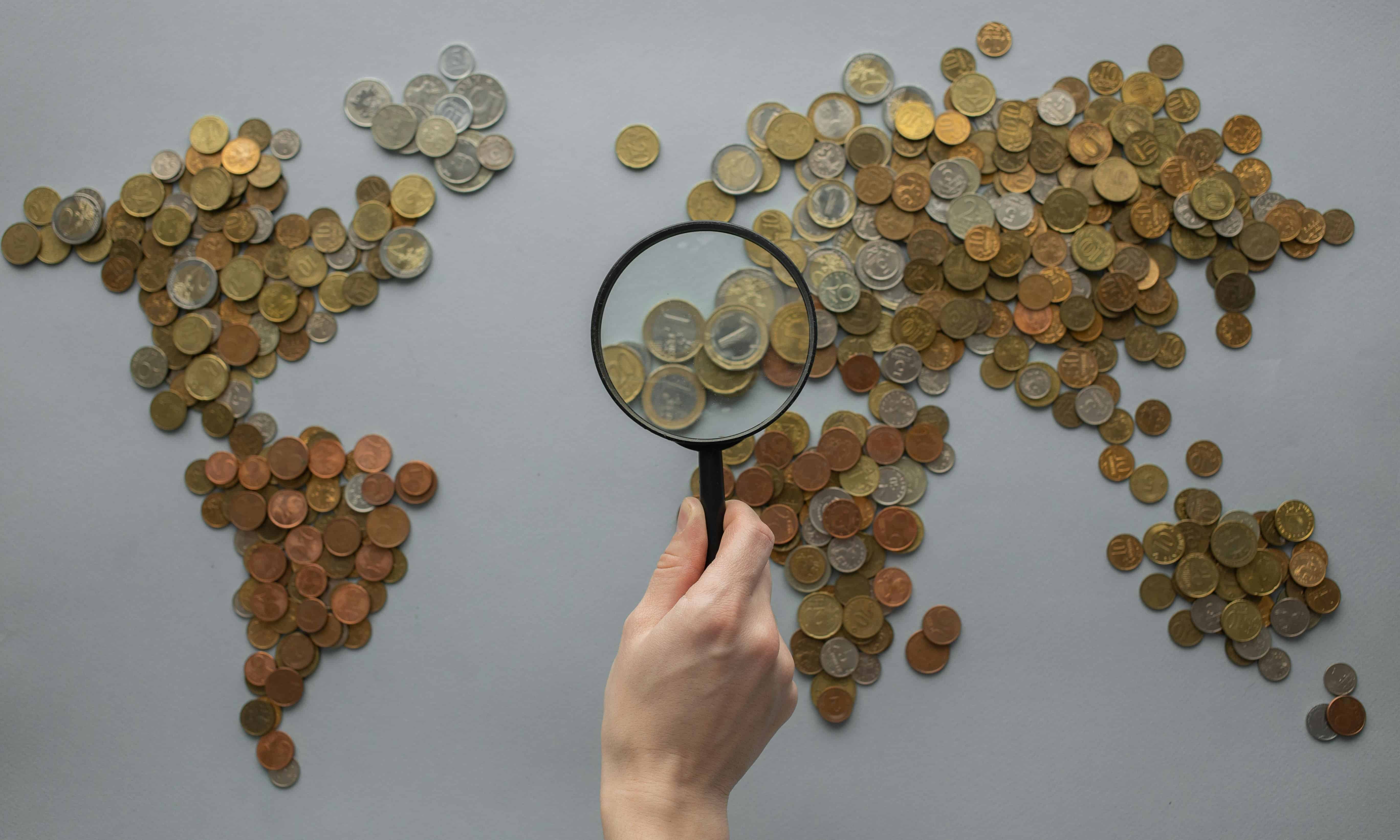 Eine Person hält eine Lupe über eine aus Münzen bestehende Weltkarte